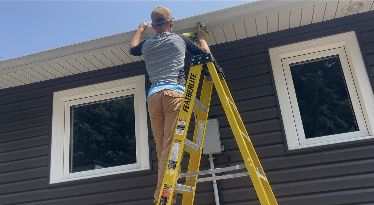 Technician on a ladder cleaning soffit and fascia along a roofline